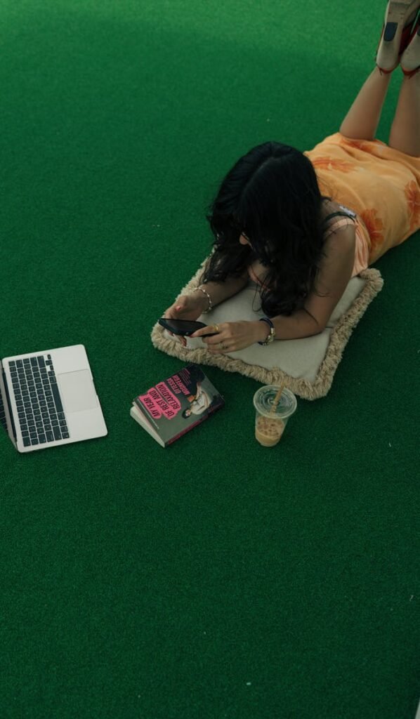 Woman using phone on artificial grass, with laptop and iced coffee nearby, creating a relaxed vibe.